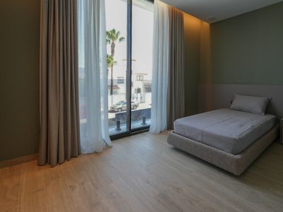 Bedroom featuring a single bed, light wood-look flooring, and olive green walls. Large windows with double curtains offer street views and palm trees.