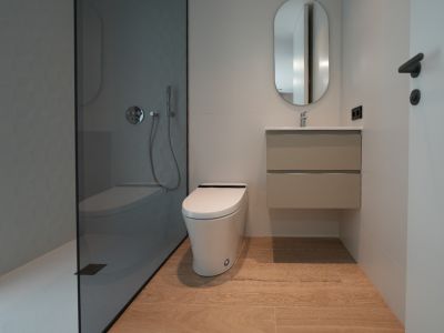 Contemporary bathroom featuring a walk-in shower with dark glass and textured grey wall, a sleek wall-mounted toilet, floating vanity, and light wood-effect floor tiles.
