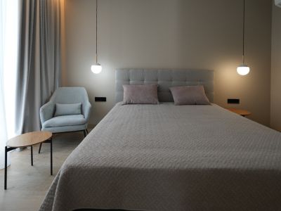 Elegant bedroom with a grey upholstered bed, two modern pendant lights, and a comfortable armchair. Light-colored curtains and wood-look flooring create a serene ambiance.