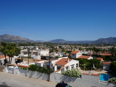 Elevated view of Albir's residential area, featuring diverse architecture, lush greenery, and a private pool, set against a backdrop of the majestic mountain range under a clear sky.