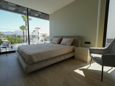 Master bedroom featuring an upholstered bed, ribbed bedside table, and expansive floor-to-ceiling windows with views of palm trees and mountains.