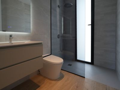Modern bathroom featuring a floating vanity, illuminated mirror, wall-mounted toilet, and a walk-in shower with rain head, all complemented by grey tiled walls and wood-effect flooring.