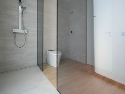 Modern bathroom featuring a walk-in shower with light grey tiles and a chrome thermostatic mixer. A glass partition separates the shower from the smart toilet area with wood-look floor tiles.