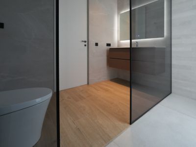 Modern bathroom featuring a wall-mounted toilet, floating wooden vanity with an illuminated mirror, and a glass shower enclosure. Wood-look floor tiles contrast with grey wall and shower tiles.