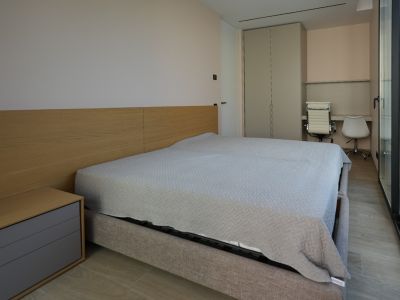 Modern bedroom featuring a light grey upholstered bed with a wooden headboard and integrated bedside table. Includes a built-in wardrobe, double desk area, and large glass doors.