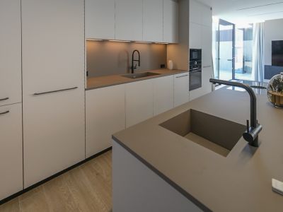 Modern kitchen featuring a central island with integrated sink, sleek flat-panel cabinetry, built-in appliances, and light wood-effect flooring, opening to an outdoor space.