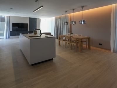 Modern kitchen island with integrated sink and hob, leading to a dining area with a wooden table, six chairs, and contemporary pendant lights. Wood-look tiled floor.