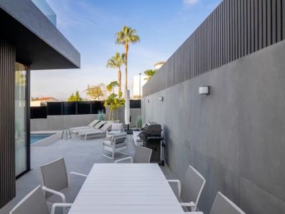 Modern outdoor terrace featuring a white slatted dining set, lounge area, and barbecue, set against grey tiled walls and dark vertical privacy screens, with a pool view.