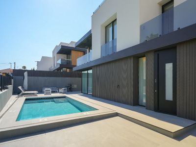 Modern villa exterior featuring a rectangular swimming pool with integrated steps, surrounded by light grey paving. The ground floor showcases a dark slatted facade and a prominent entrance door.