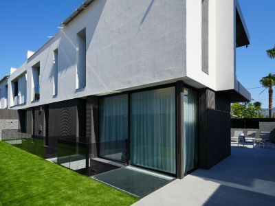 Modern villa exterior showcasing contrasting white/grey and dark textured facades, large glass doors, a manicured lawn, and a tiled patio with outdoor seating.