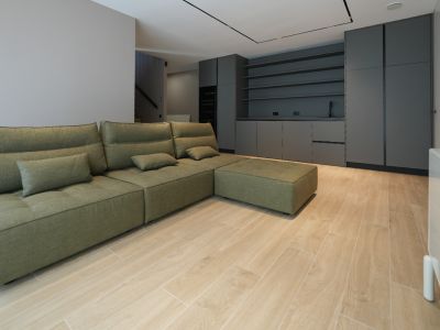 Olive green modular sofa in an open-plan living space with light wood-look tile flooring. Dark grey built-in cabinetry with a sink and shelving visible in the background.