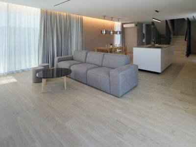 Open-plan living room featuring a grey modular sofa, black marble coffee table, and wood-look tiled floor. Large windows with sheer and grey curtains.