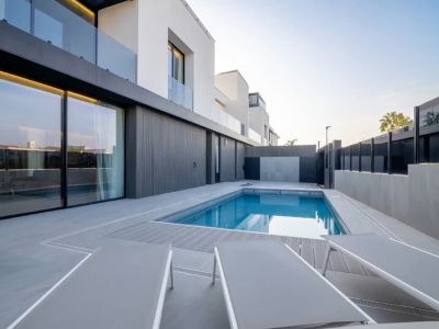 Outdoor pool with blue water, bordered by a grey tiled patio and composite decking. Modern facade with large sliding glass doors reflects the clear sky.