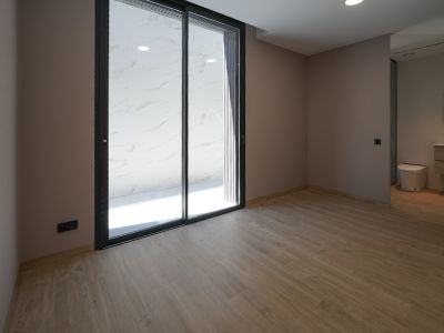 Spacious room with wood-look tiled flooring and light-colored walls. Large black-framed sliding glass doors open to an outdoor area with white marble-patterned tiles. Integrated bathroom visible.