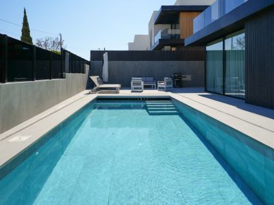 The outdoor area features a rectangular pool with light blue tiles and steps, surrounded by light grey paving. A modern lounge set and sun lounger offer relaxation beside the sleek facade.