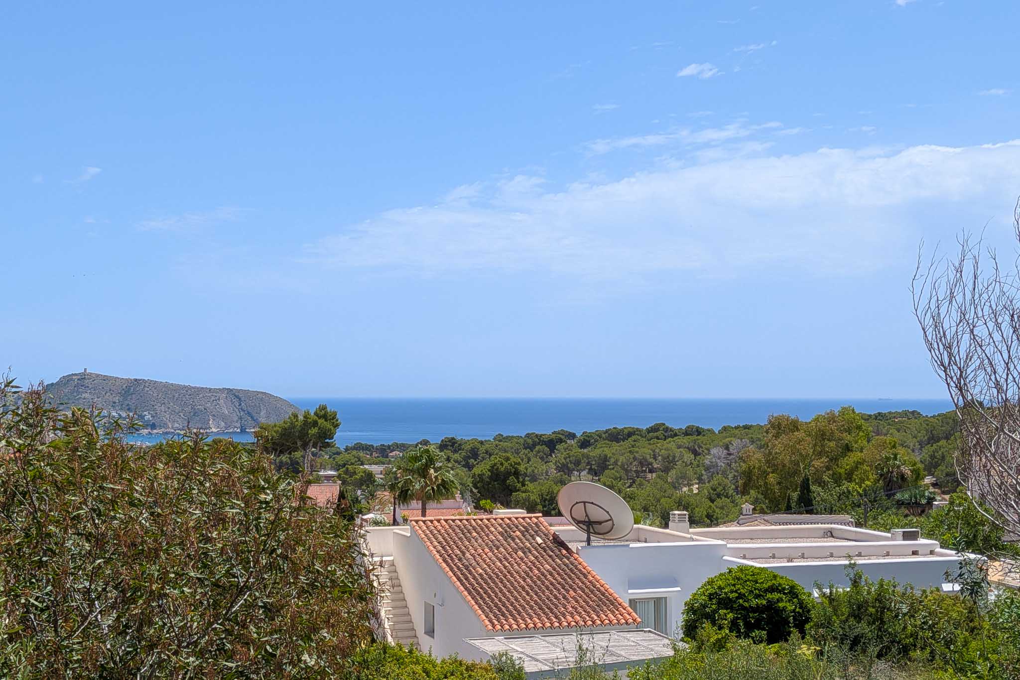 Elevated sea view of the Mediterranean, featuring traditional terracotta roofs and lush foreground vegetation. A rocky headland with a distant tower stands against a clear blue sky.
