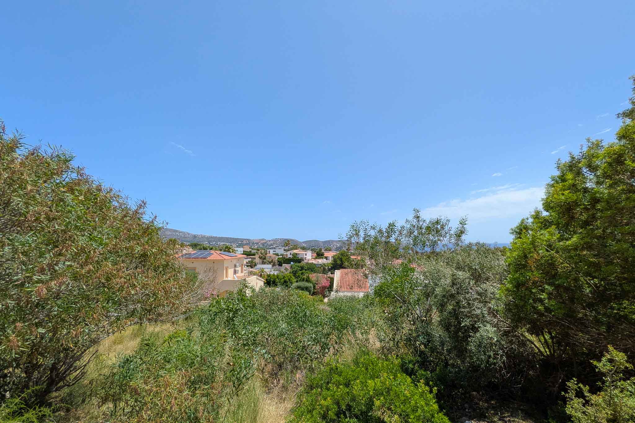 Panoramic view from the plot, revealing a green residential area with traditional rooftops, distant sea, and mountains under a clear blue sky.