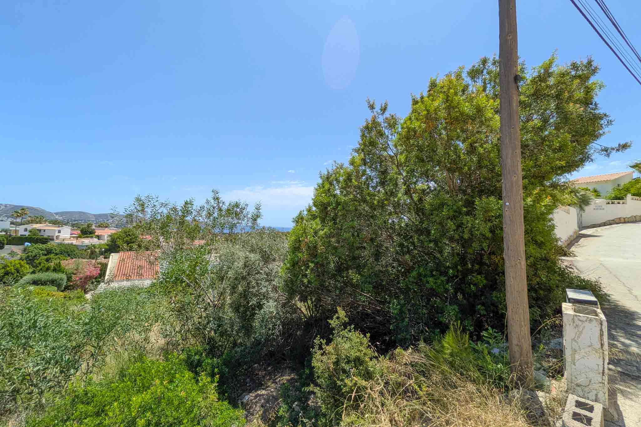 Undeveloped plot with dense Mediterranean vegetation, a utility pole, and a paved road. Offers distant views of residential rooftops, hills, and a clear blue sky with a glimpse of the sea.