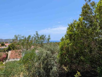 Panoramic view from the plot, featuring lush Mediterranean vegetation in the foreground, residential rooftops, and the distant Mediterranean Sea under a clear blue sky.