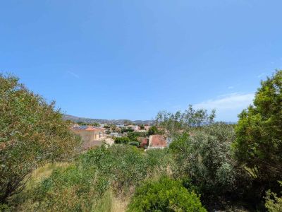 Panoramic view from the plot, revealing a green residential area with traditional rooftops, distant sea, and mountains under a clear blue sky.