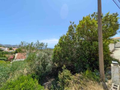 Undeveloped plot with dense Mediterranean vegetation, a utility pole, and a paved road. Offers distant views of residential rooftops, hills, and a clear blue sky with a glimpse of the sea.