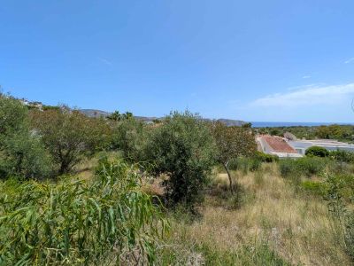 Undeveloped plot with natural Mediterranean vegetation, offering partial sea views framed by residential rooftops and distant hills under a clear blue sky.