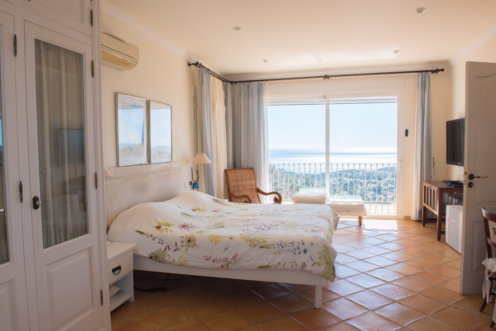 Bright bedroom featuring terracotta tile flooring, built-in white wardrobes, and sliding glass doors opening to a balcony with expansive sea views.