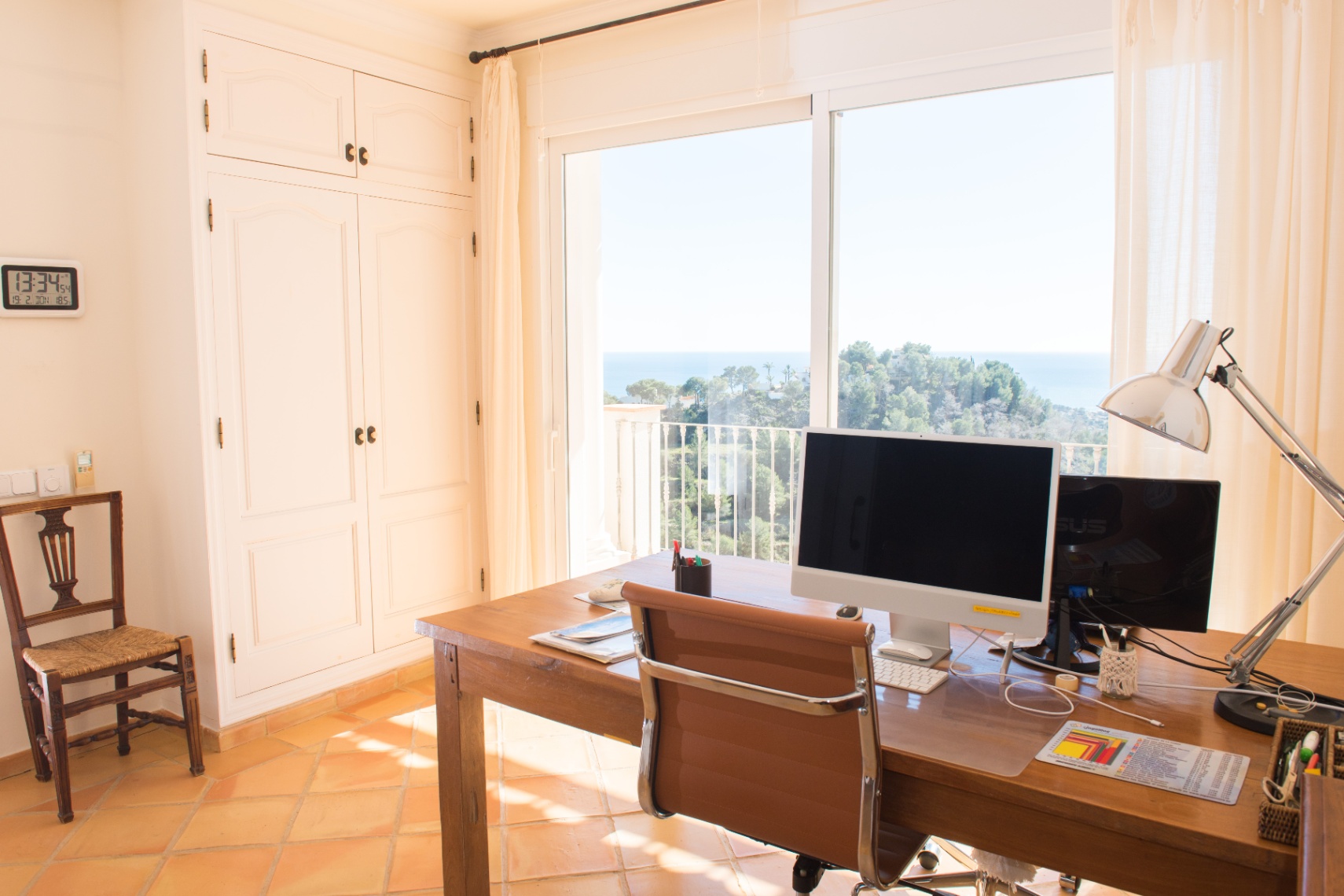 Bright home office featuring a large wooden desk, built-in white wardrobes, and floor-to-ceiling windows with panoramic sea and coastal views.