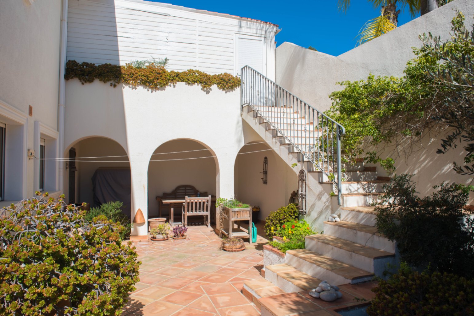 Exterior courtyard with terracotta tiling, featuring arched shaded alcoves and a stone staircase with metal railing leading to the upper floor.