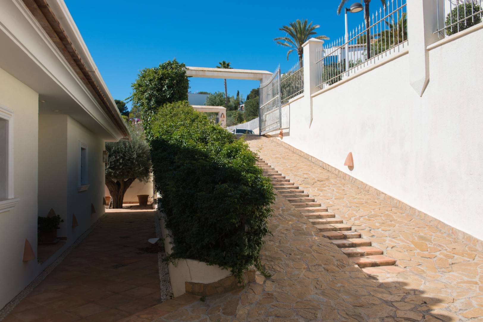 Exterior entrance featuring a natural stone-paved driveway with integrated steps, flanked by white perimeter walls and manicured Mediterranean hedges.