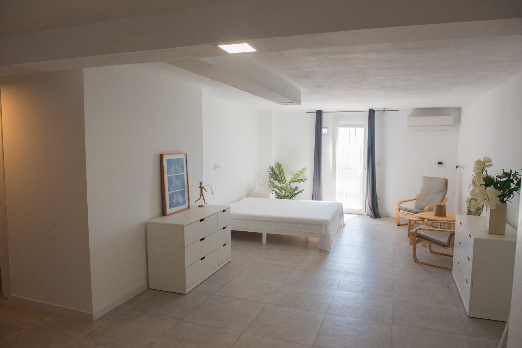 Minimalist bedroom with neutral tones, grey ceramic flooring, functional white furniture, air conditioning, and direct outdoor access.