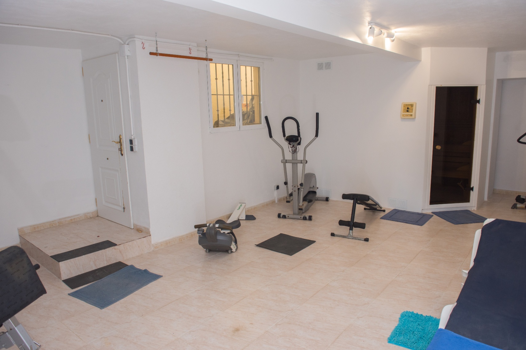 Private home gym equipped with cardio machines, a weight bench, and direct access to a sauna. The space features neutral tiled flooring.
