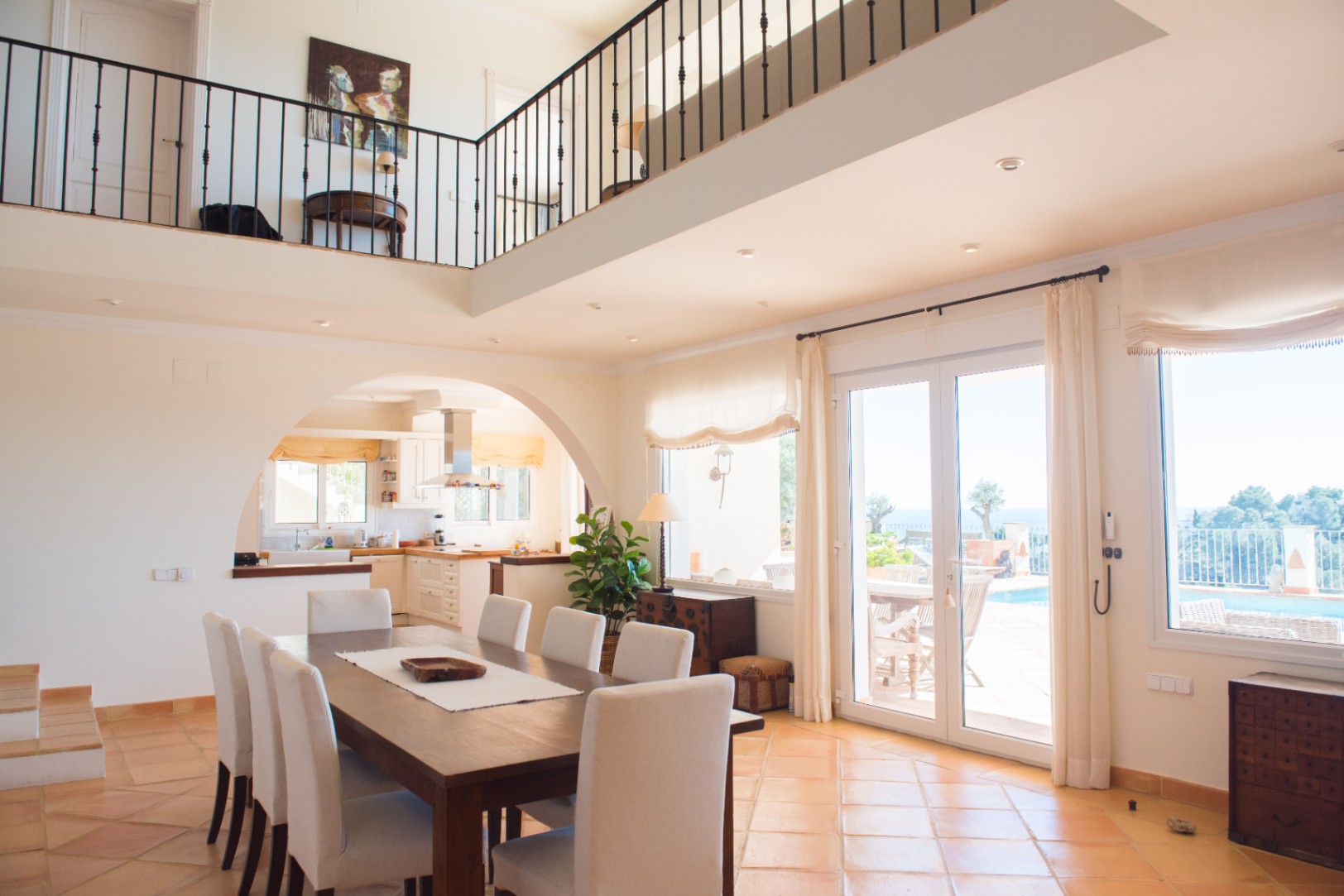 Spacious dining area featuring terracotta tile flooring, a large wooden table, and an arched entrance to the kitchen. A mezzanine gallery overlooks the bright space.