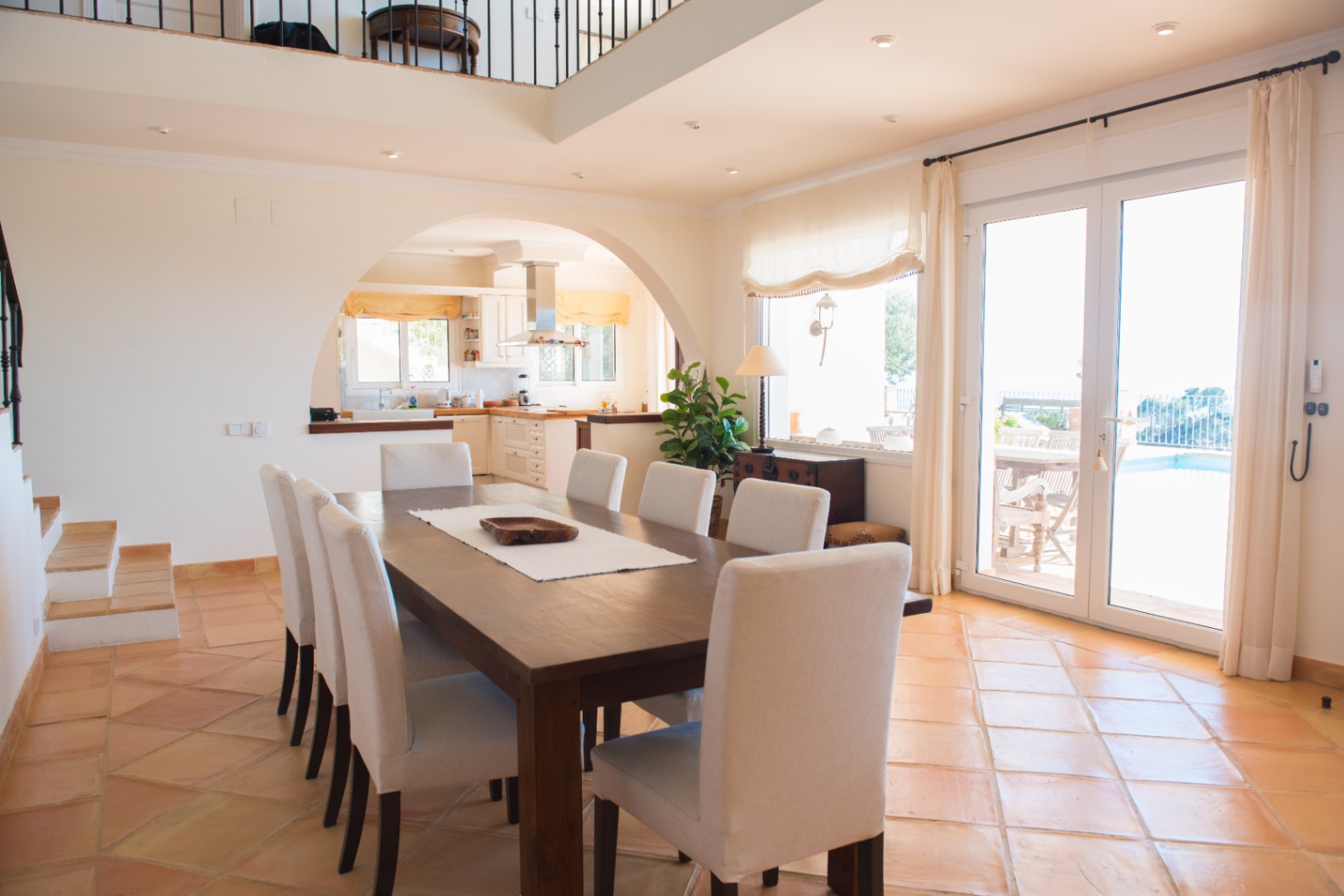 Spacious dining room featuring a solid wood table, terracotta tile flooring, and an arched opening to the kitchen, with direct access to the pool terrace.