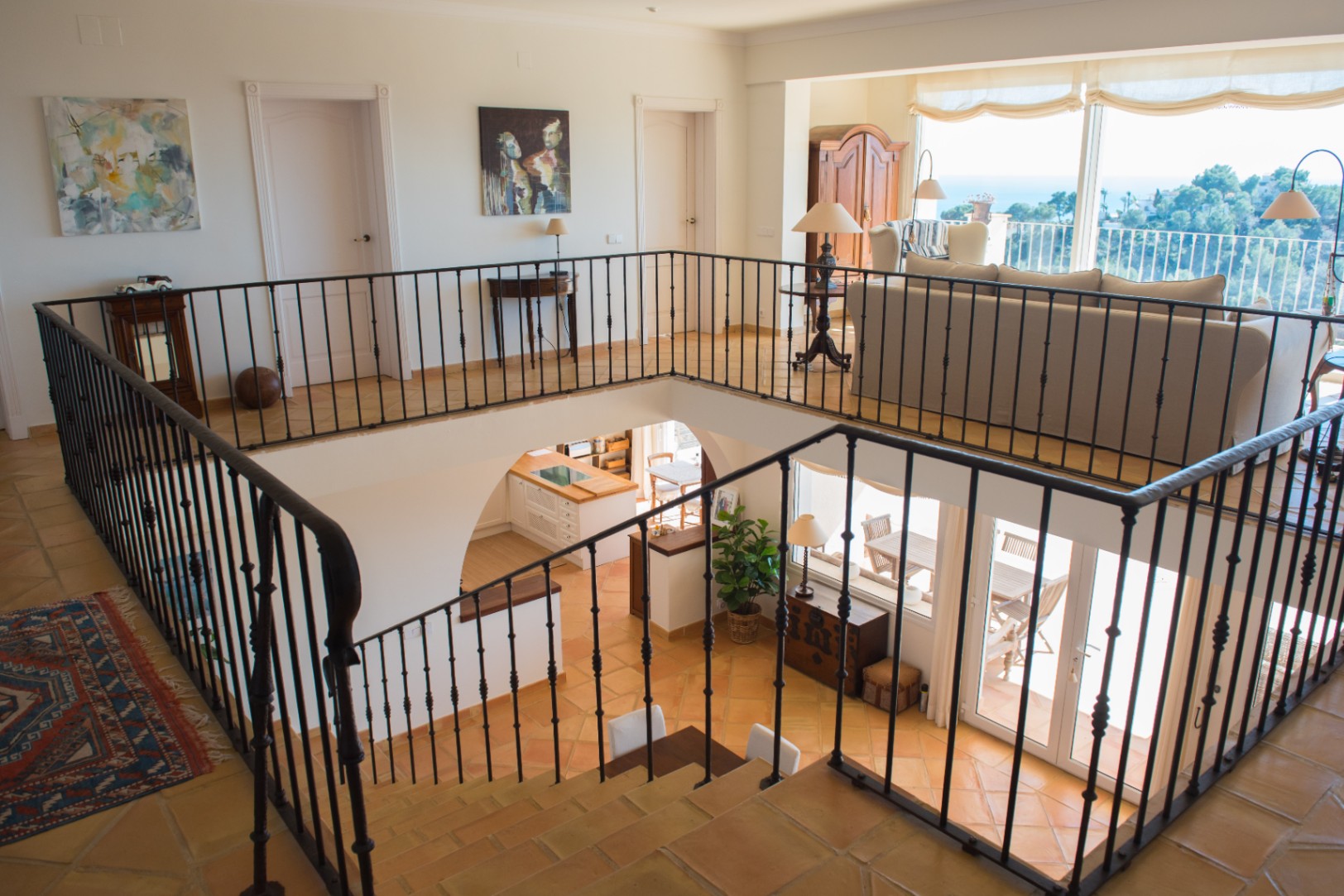 Spacious landing with wrought iron railings and terracotta tiles, overlooking the lower living area and offering panoramic sea views through large windows.