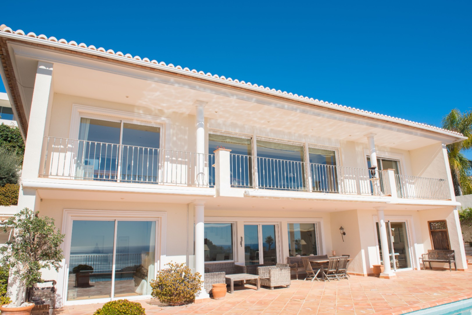 Two-story villa exterior featuring white columns, terracotta tiling, and expansive sliding glass doors leading to a spacious terrace and balcony.