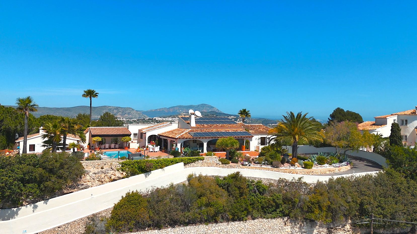 Aerial view of the villa featuring terracotta roofs with solar panels, a private pool, and landscaped gardens overlooking the Mediterranean coast.