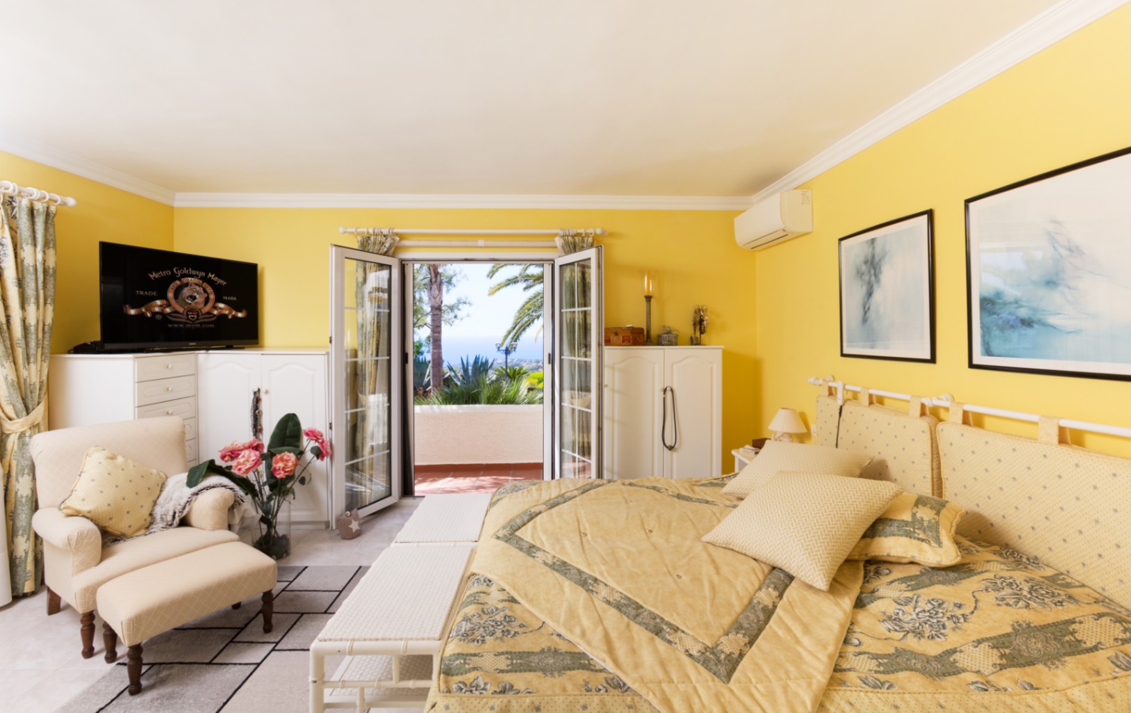 Bedroom featuring yellow walls, white cabinetry, and a seating area. Double glass doors open to a private terrace with sea views, palm trees, and Mediterranean plants.