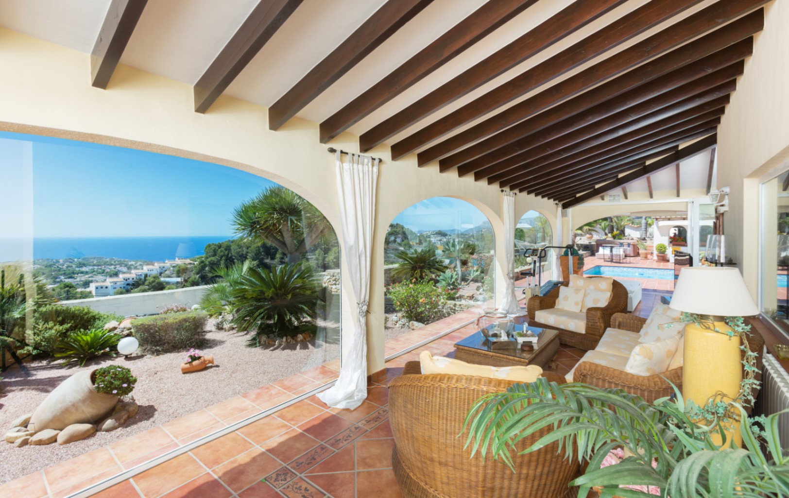 Covered terrace featuring dark wood ceiling beams, terracotta flooring, and wicker seating. Arched glass enclosures provide panoramic sea views and garden access.