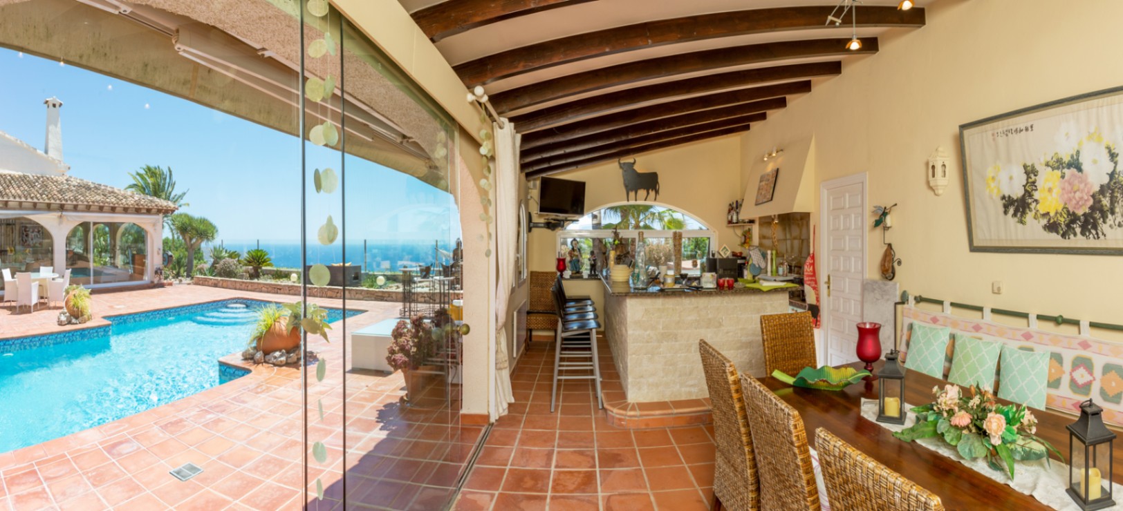 Covered terrace featuring exposed wooden beams, terracotta tiling, and an outdoor stone bar, overlooking the swimming pool and panoramic sea views.