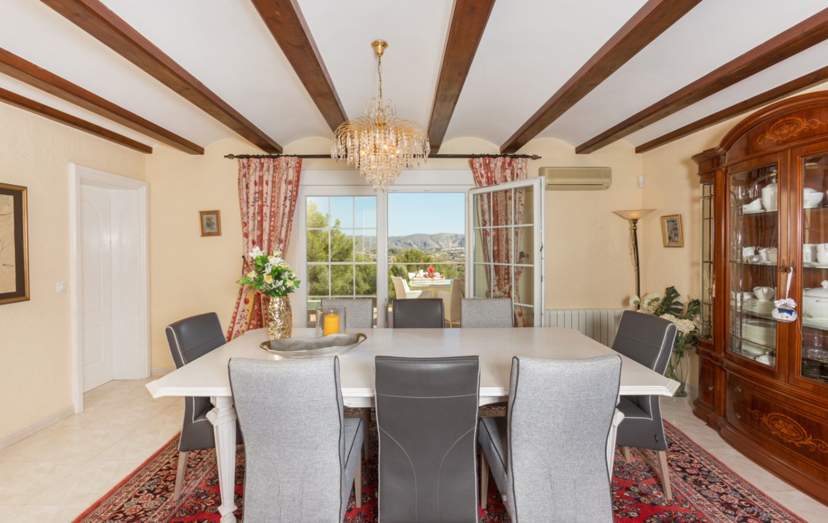 Dining room featuring exposed wooden ceiling beams, a white table with grey chairs, and a classic display cabinet. Large doors open to a terrace with mountain views.