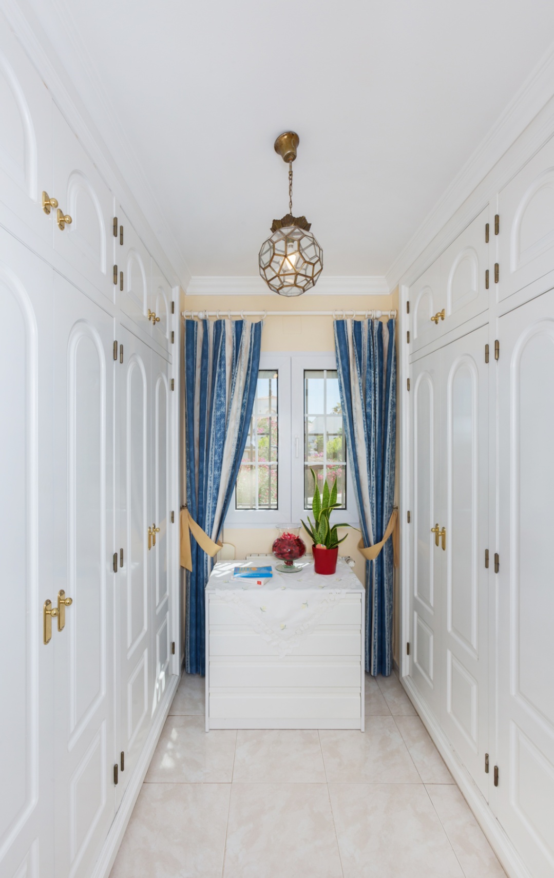 Dressing area featuring floor-to-ceiling white wardrobes with arched paneling, brass hardware, a central window with blue curtains, and a geometric pendant light.