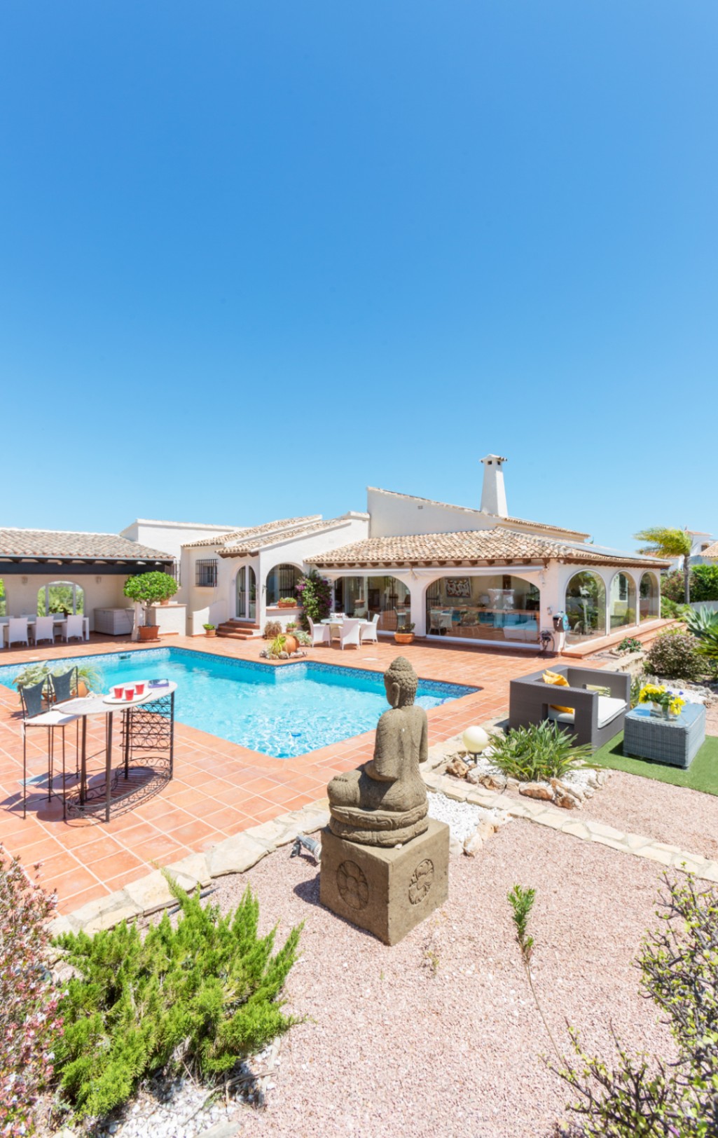 Exterior terrace with a blue-tiled swimming pool, terracotta flooring, a stone Buddha statue, and a Mediterranean villa featuring a glass-enclosed porch.