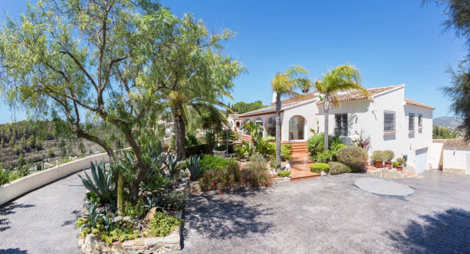 Exterior view of the Mediterranean villa featuring a paved driveway, landscaped gardens with agaves and palm trees, and a white facade with arched details.