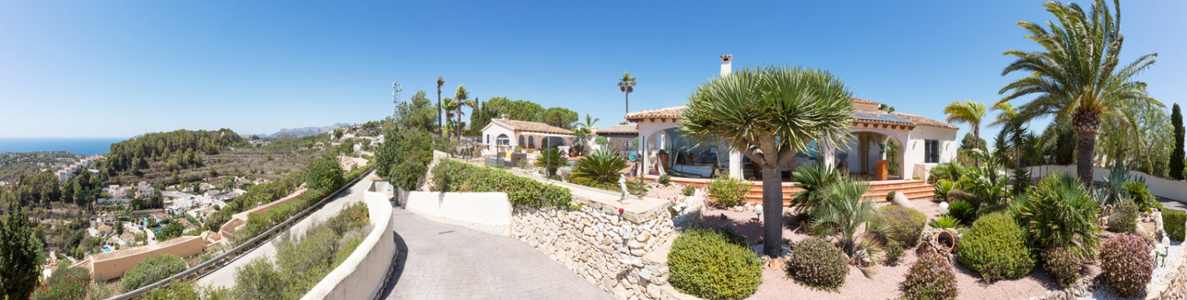 Panoramic view of the villa's exterior featuring a paved driveway, traditional stone walls, and a landscaped Mediterranean garden with palm trees overlooking the coast.