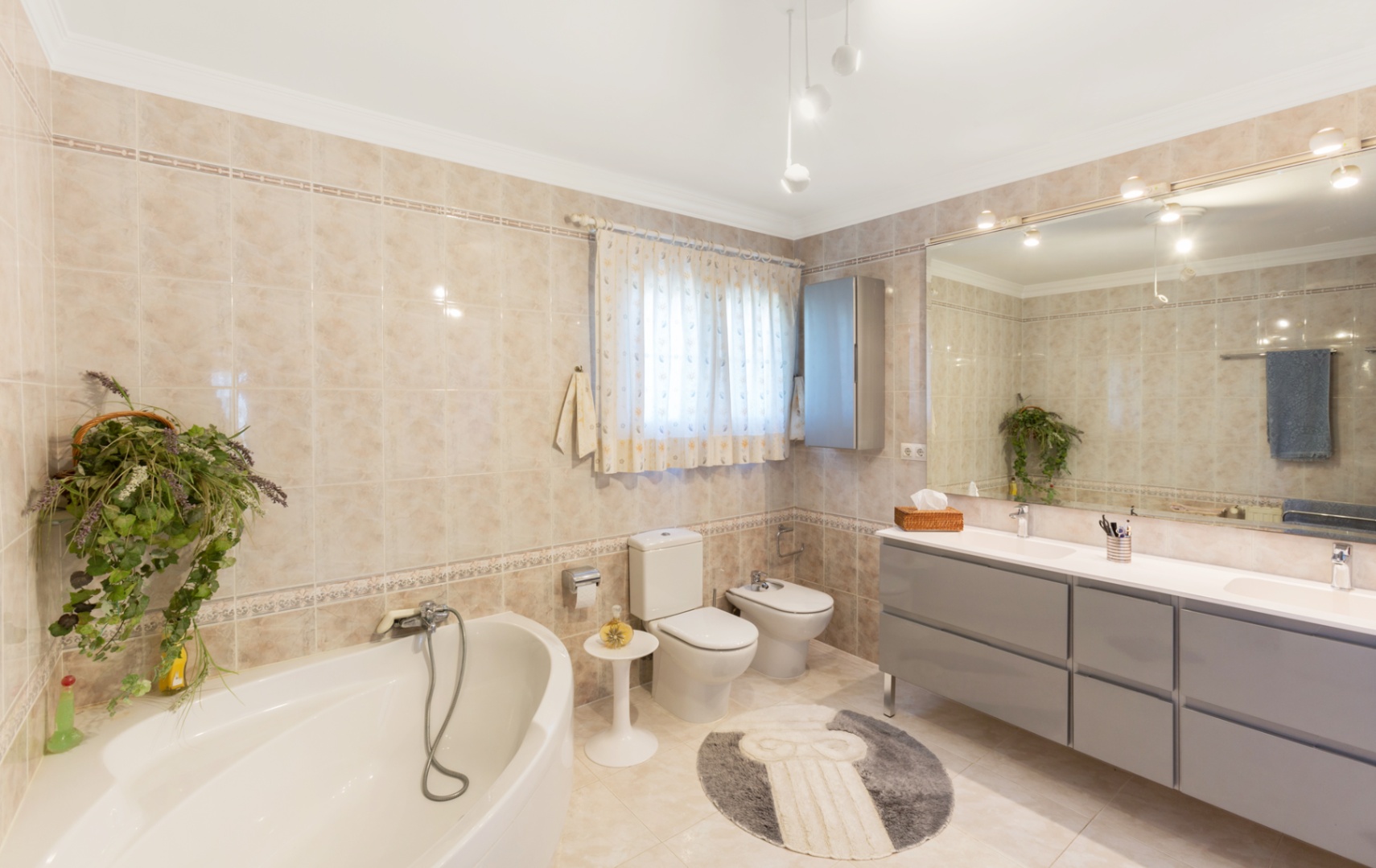 Spacious bathroom featuring a corner bathtub, a double vanity with grey cabinetry, and a large wall mirror. Beige tiling with decorative borders covers the surfaces.
