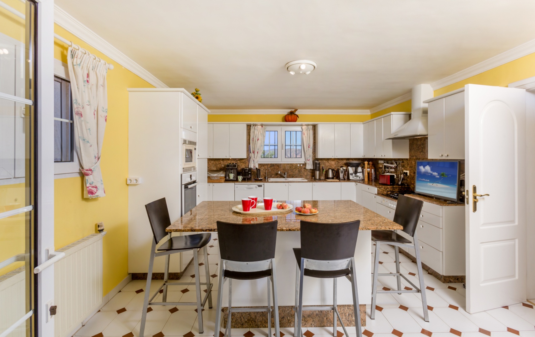 Spacious kitchen featuring a granite island with seating for four, white cabinetry, yellow walls, and integrated appliances. The tiled floor includes decorative brown accents.