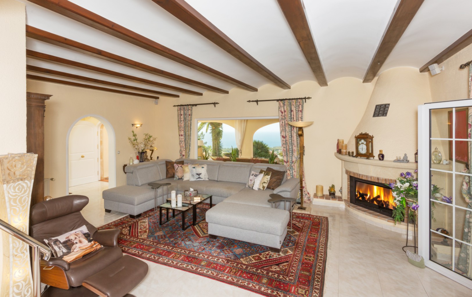 Spacious living room featuring exposed wooden ceiling beams, a large grey sectional sofa, and a traditional fireplace with sea views.