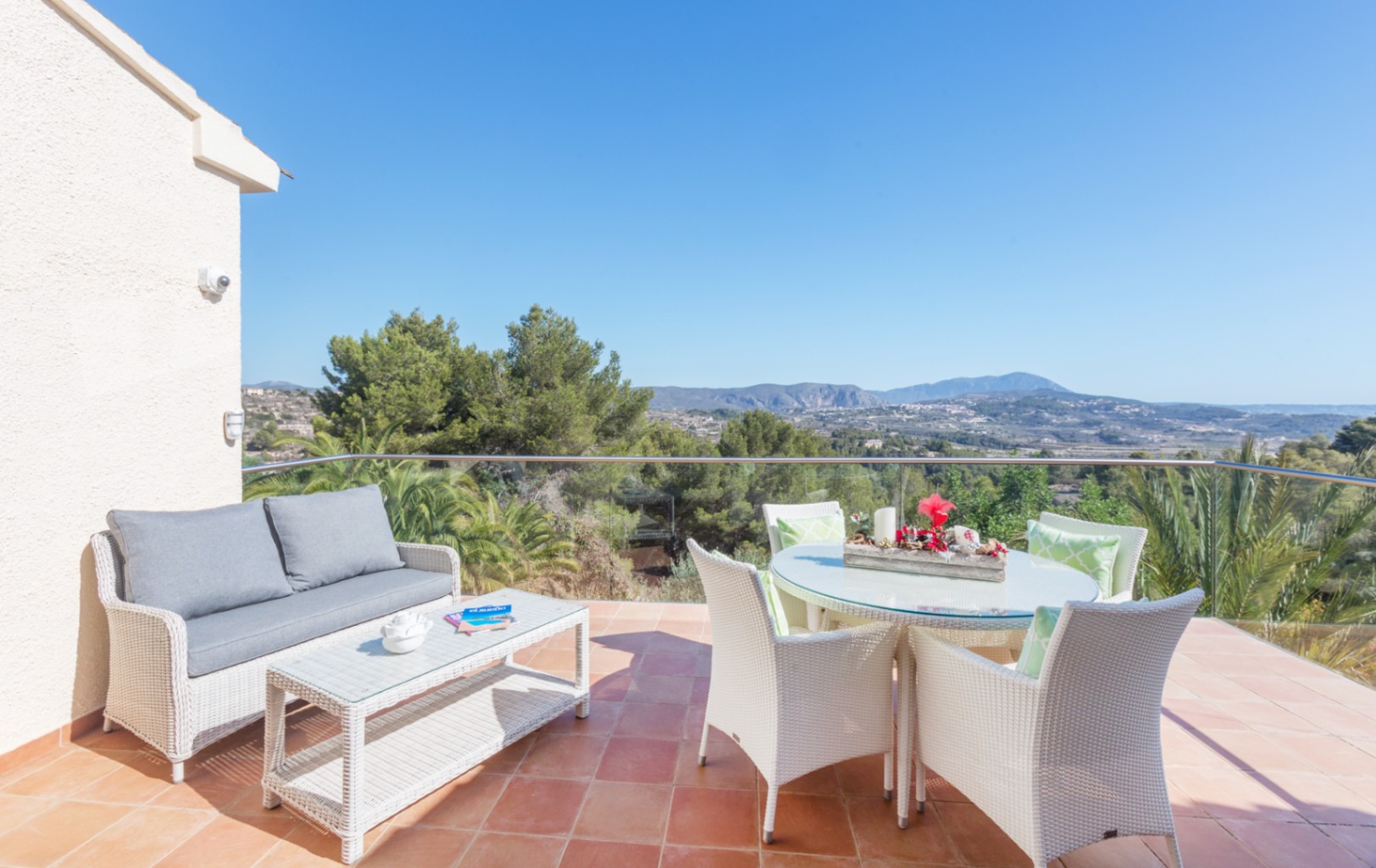 Terrace with terracotta tiles featuring a white wicker lounge and dining set, enclosed by glass railings with views of the surrounding mountains and pine trees.