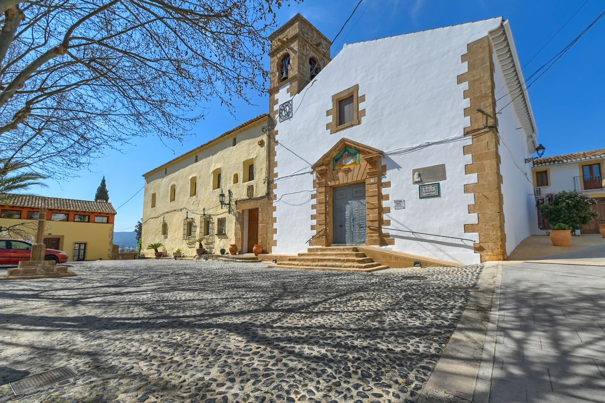 Cobblestone church square in Jesús Pobre featuring a historic white facade with stone accents, a bell tower, and traditional Mediterranean architecture.